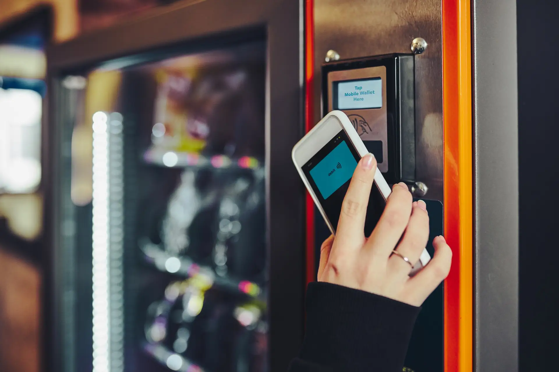 Hand hält ein Smartphone zum kontaktlosen Bezahlen an das Kartenterminal eines Verkaufsautomaten