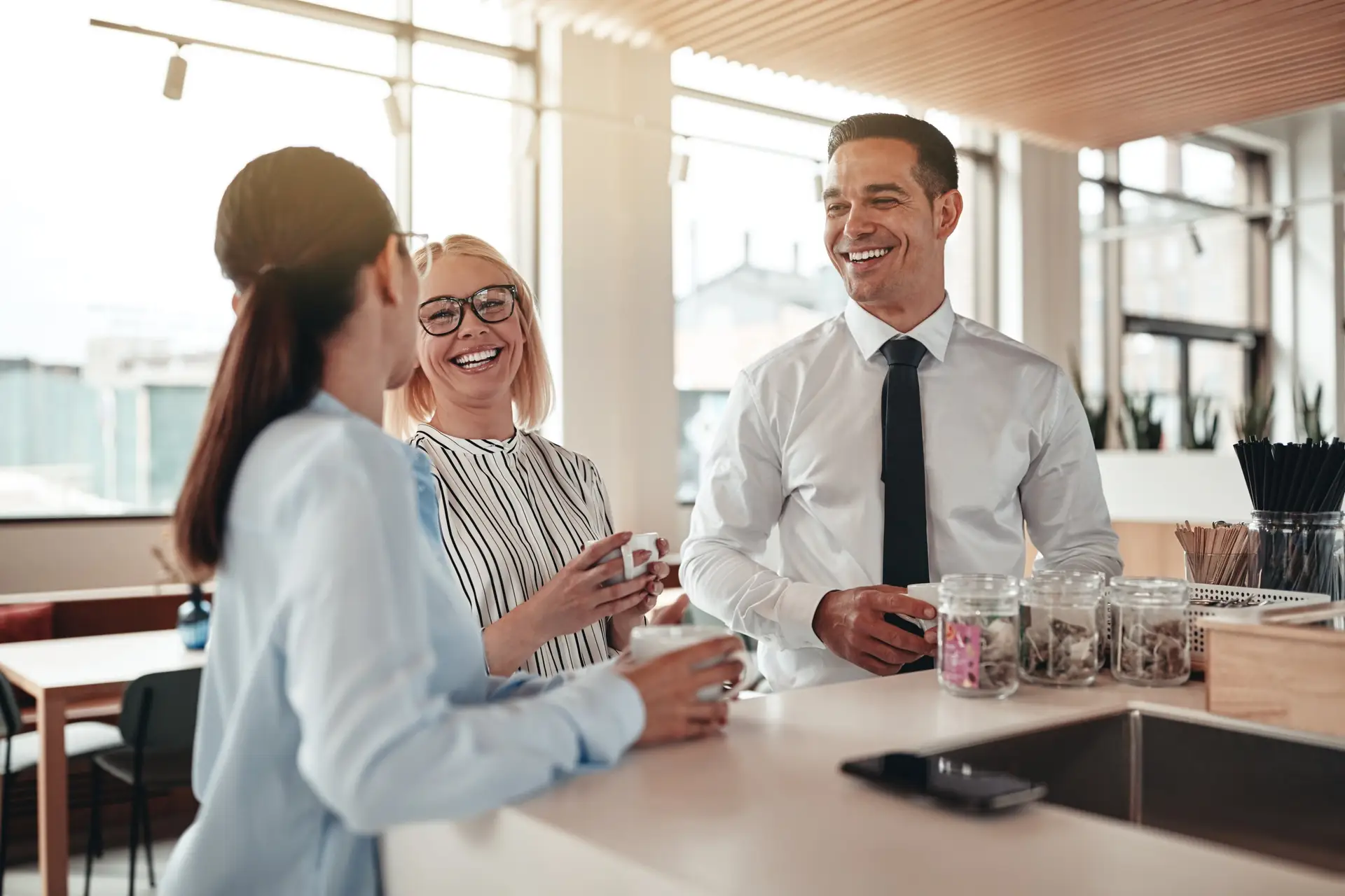 Drei Kolleginnen und Kollegen trinken lachend Kaffee an der Theke der Büroküche