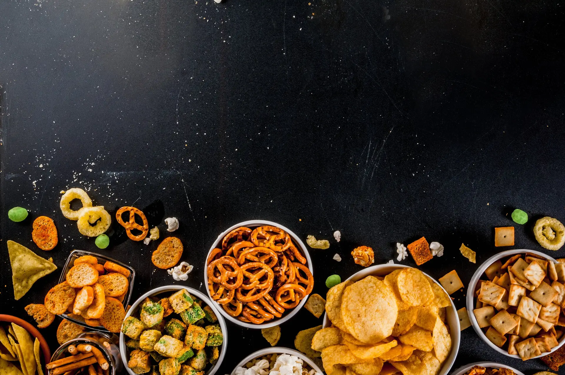 Brezeln, Chips und Cracker in Schalen auf dunklem Untergrund